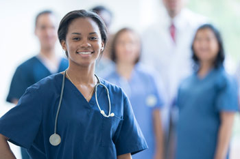 A black woman doctor wearing blue scrubs and a stethoscope smiles. Her colleagues are standing behind her, out of the camera's focus.