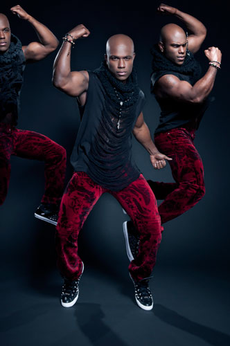 Milan Christopher poses in red pants and a black shirt, flexing the muscles of his bare arms and balancing on the points of his shoes.