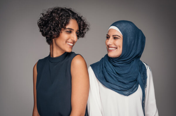 Two women looking at one another and smiling. One is wearing a black sleeveless blouse and the other is wearing a white long sleeve shirt and a blue headscarf