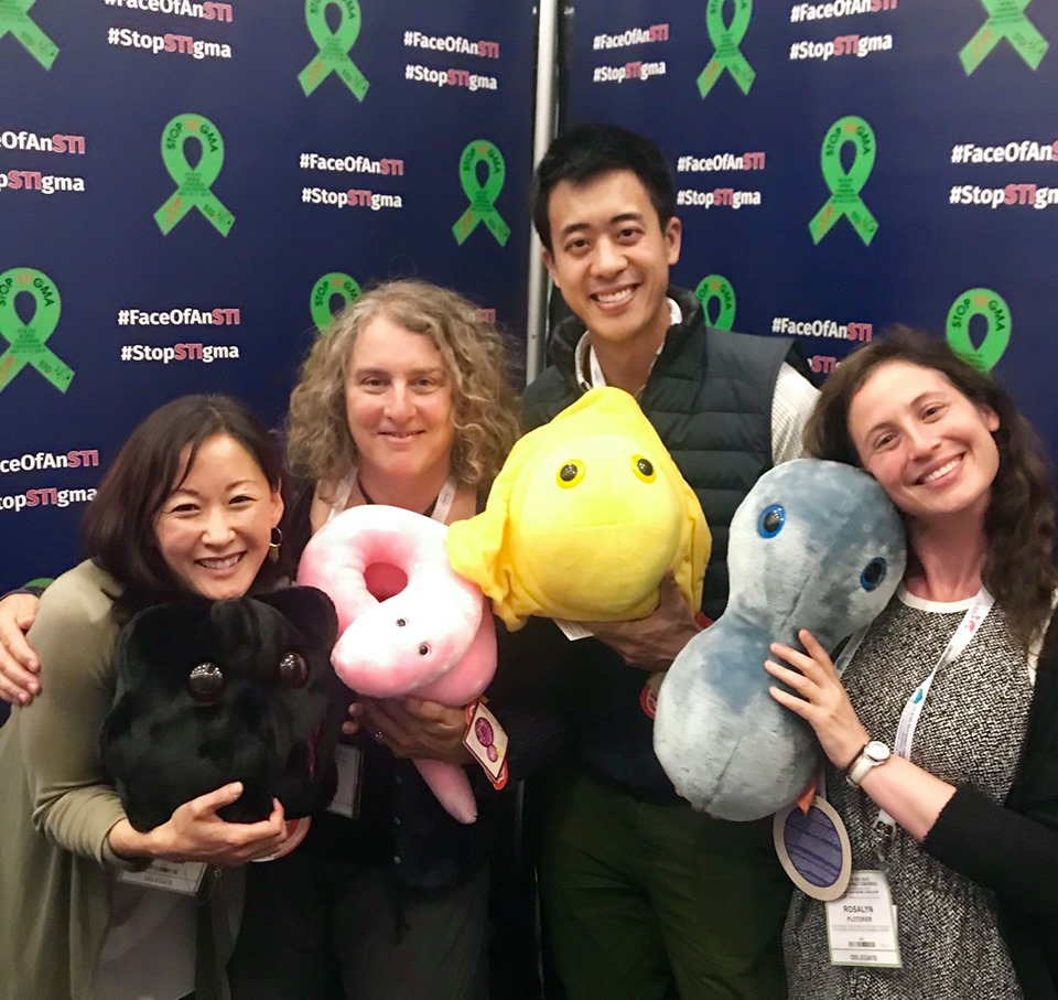 Doctors Ina Park, Sharon Adler, Eric Tang, and Rosalyn Plotzker hold up plush stuffed toys of HIV, syphilis, herpes and gonorrhea microbes