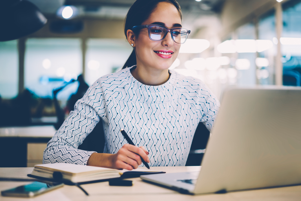 Person with glasses at their laptop smiling and taking notes.