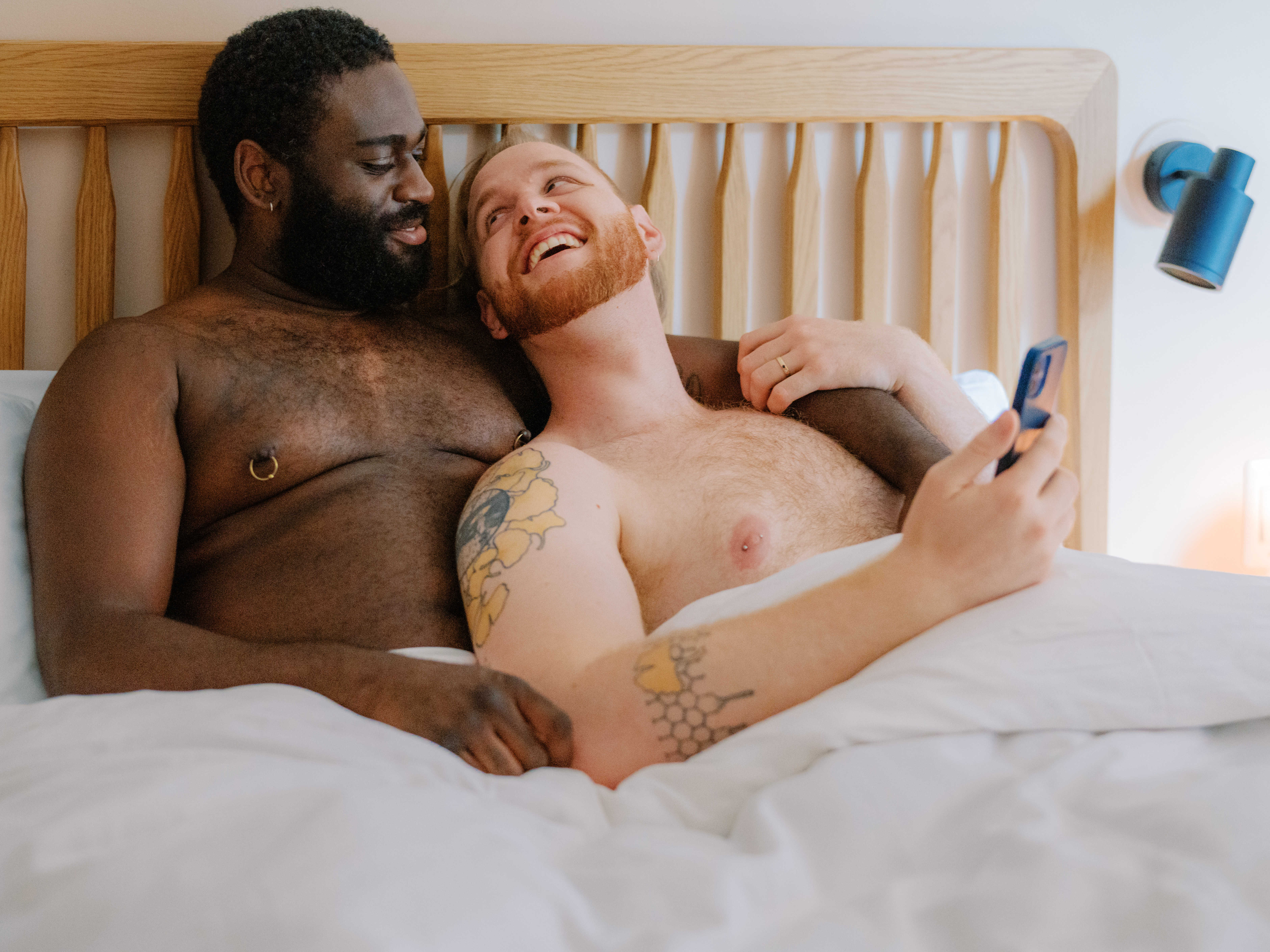A queer couple uses a phone in bed together.